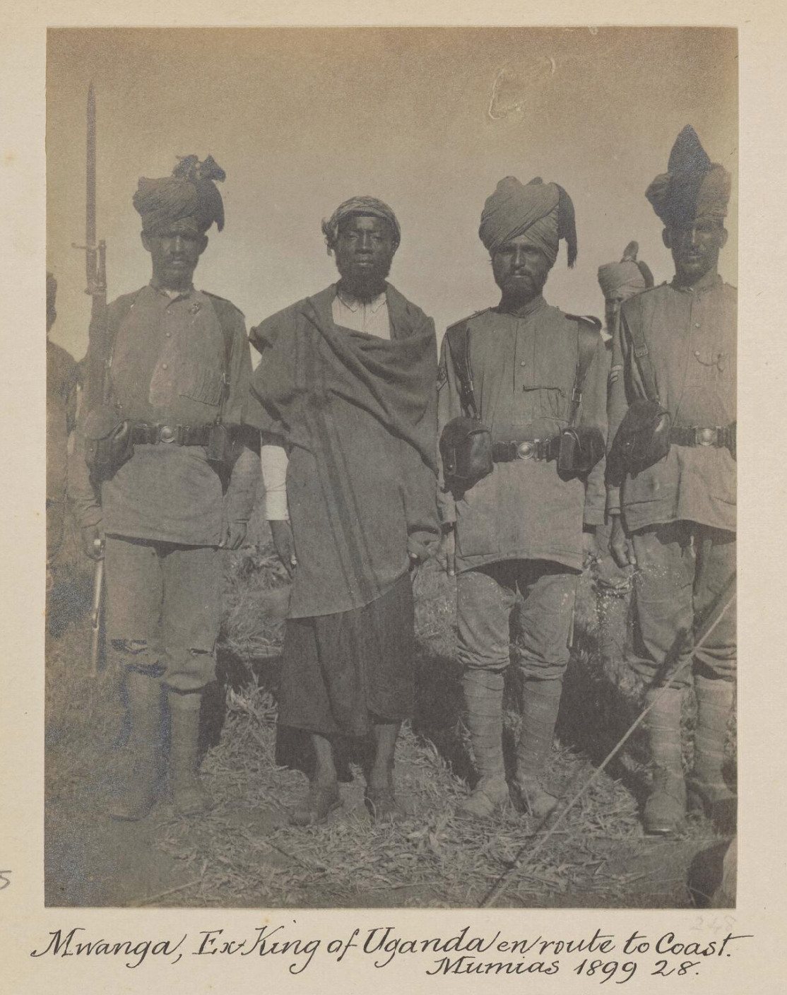 Mwanga, wrapped in a dark cloth with a head wrap, stands between three turbaned, uniformed soldiers with rifles; handwritten caption below: Mwanga, Ex-King of Uganda en route to Coast, Mumias 1899.