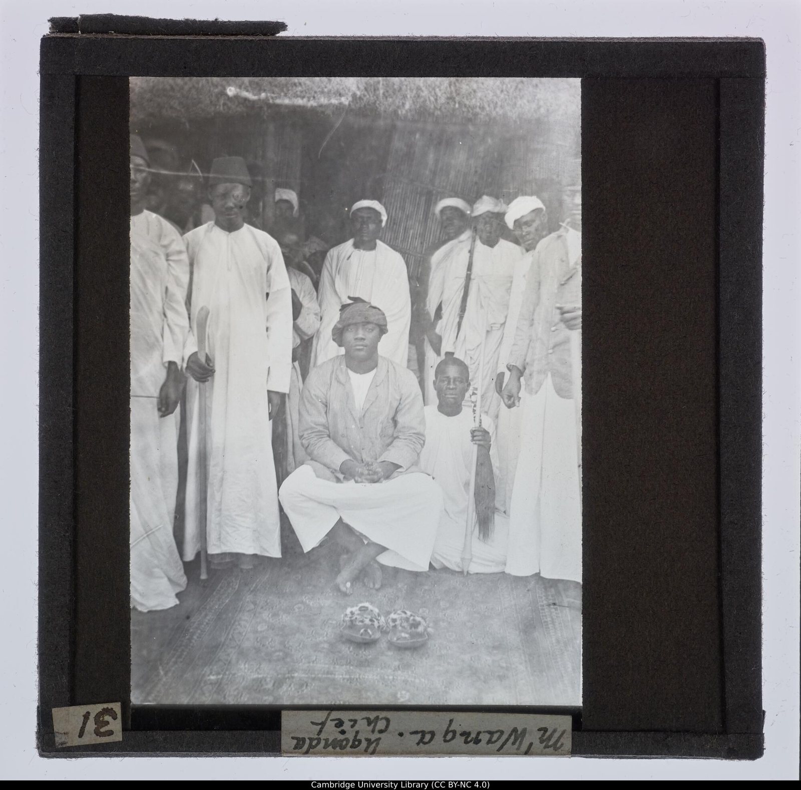 Glass lantern slide: Kabaka Mwanga sits cross-legged at the centre of a group of chiefs in white robes, a boy holding a staff beside him; the slide's label reads M'Wanga Uganda Chief.
