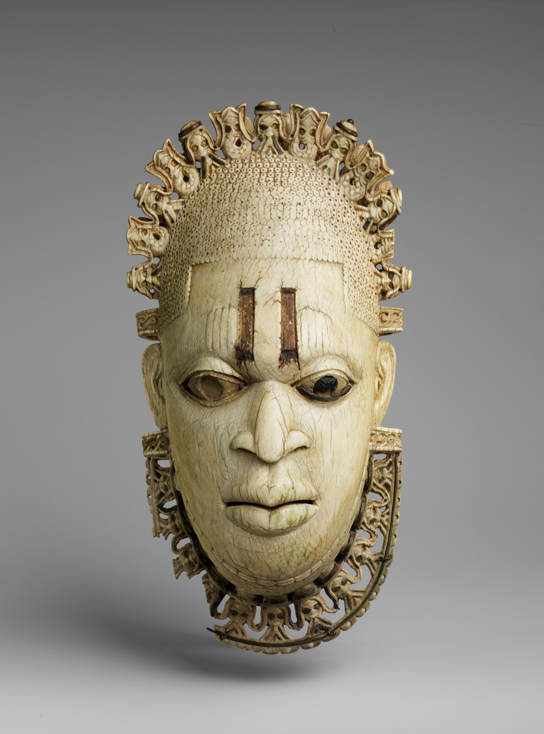 Ivory pendant mask of a woman’s face with two dark vertical marks on the forehead and inlaid eyes, framed above by an openwork border of small Portuguese heads alternating with stylised mudfish, and below by a similar collar.