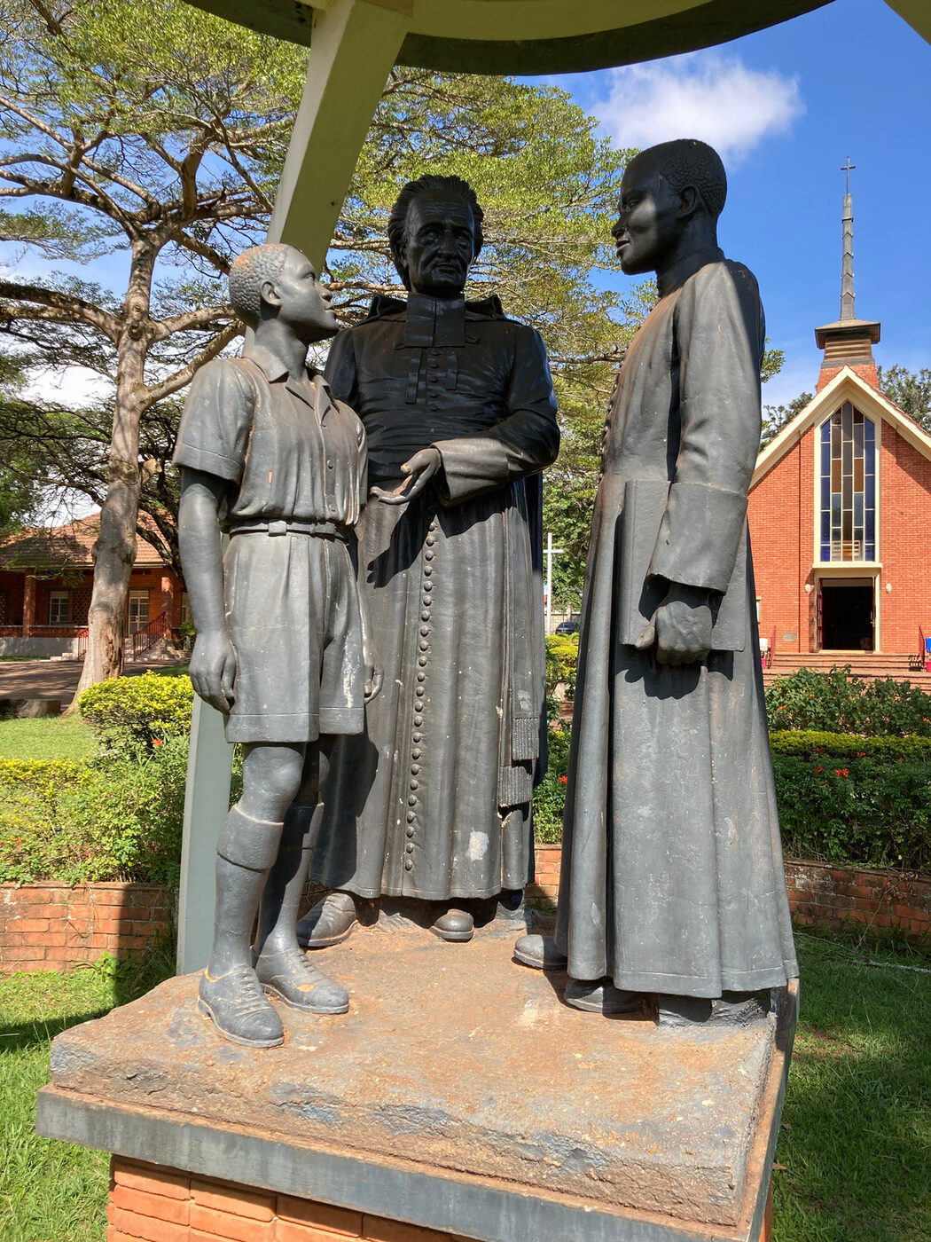Three life-size concrete figures on a brick plinth under a curved canopy: a boy in shorts, a priest, and a young man in a cassock, with a red-brick church behind.