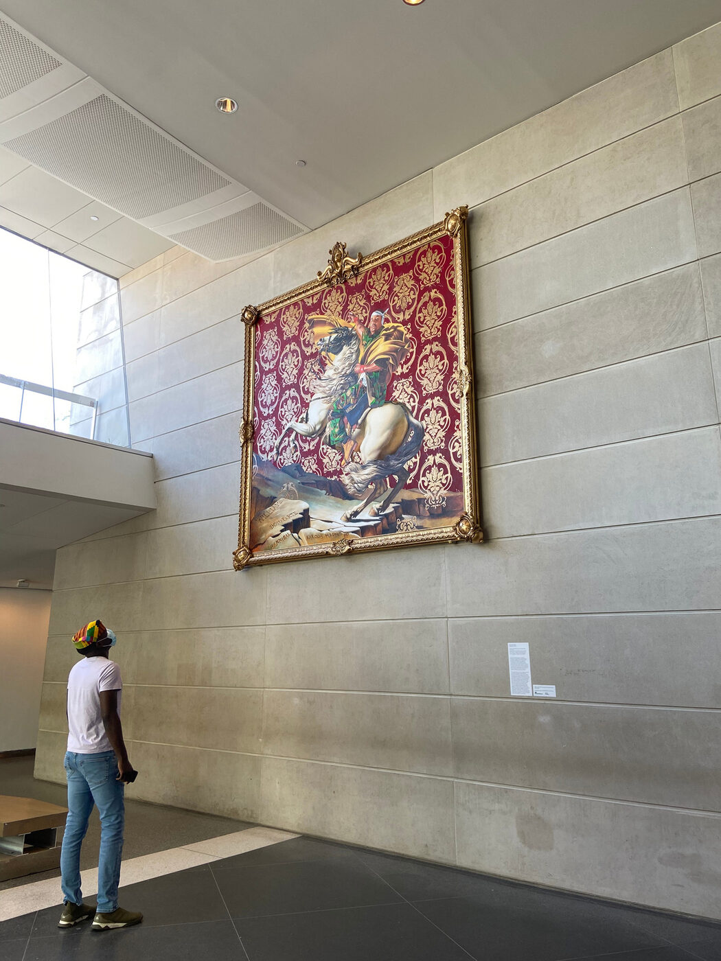 Mikka, small in the frame, looking up at Kehinde Wiley's large painting in a gilded frame on a pale museum wall.