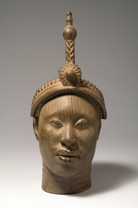 A cast copper-alloy head with a tall beaded crown and fine vertical facial striations, on a pale ground.
