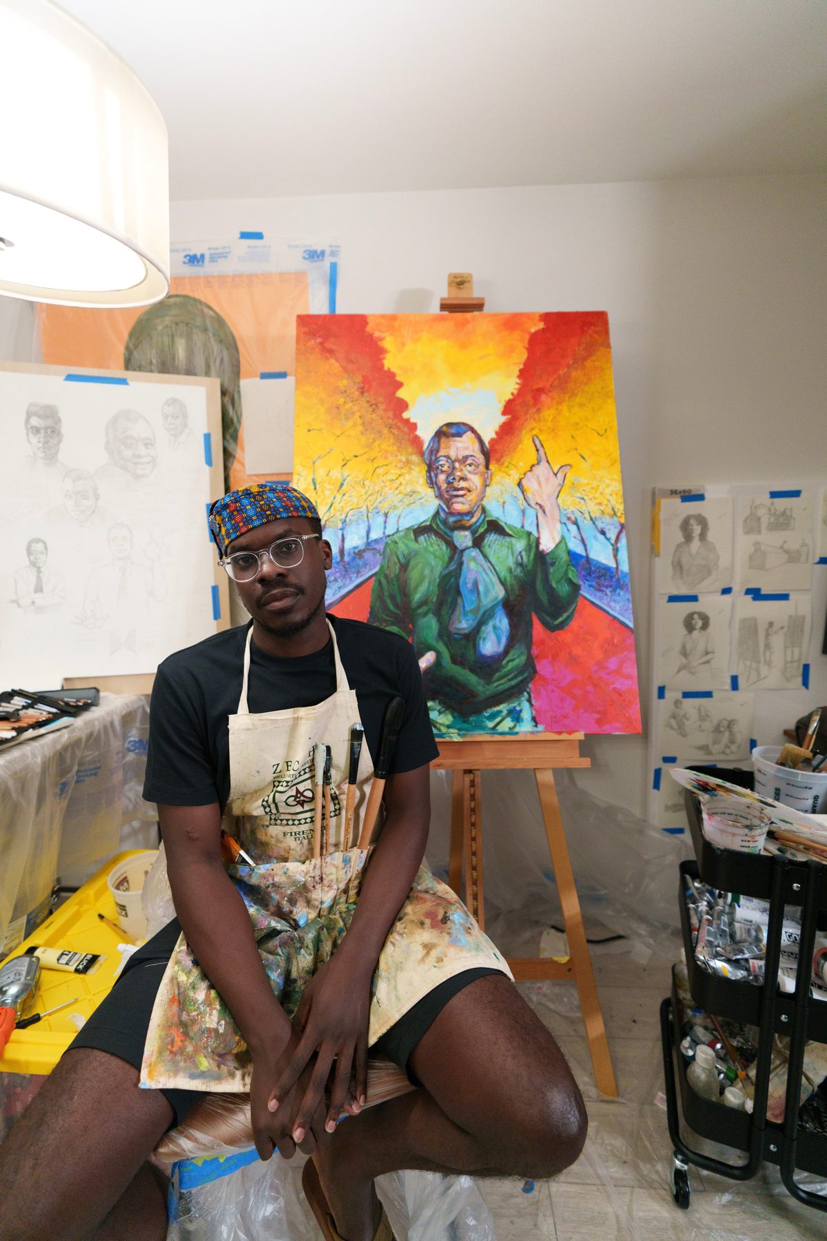 Mikka Kabugo seated in front of the Baldwin portrait, brushes in apron pocket.